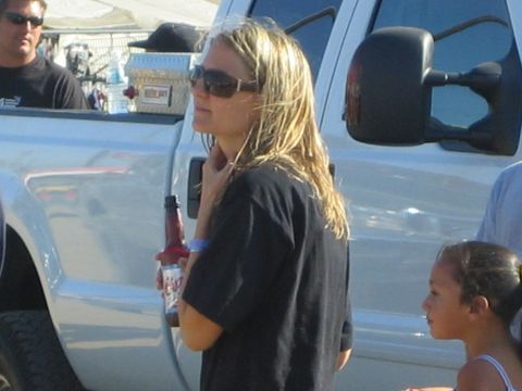 1990 Ford Bronco - woman in sunglasses and a dark T-shirt holds a beer bottle beside a white pickup, with a child and a cooler nearby in a dusty lot.
