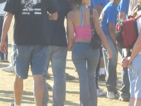 1990 Ford Bronco - woman in a pink halter top and jeans with a shoulder bag walks away through a crowd of people in a dirt lot.