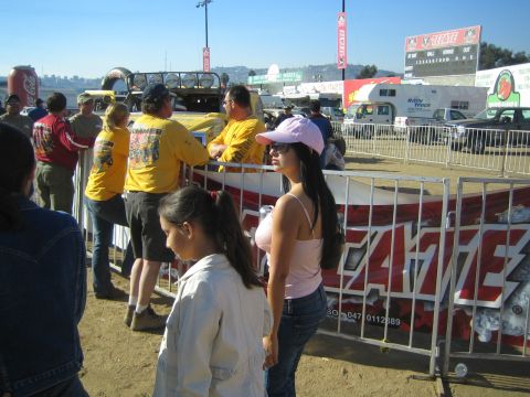 1990 Ford Bronco - woman in a pink visor and tank top and a girl stand by a red Tecate barricade as crew in yellow shirts gather near a grandstand.