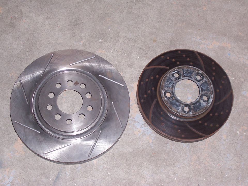 Two brake rotors on a concrete surface; one is new with slots, the other is old and rusted with drilled holes