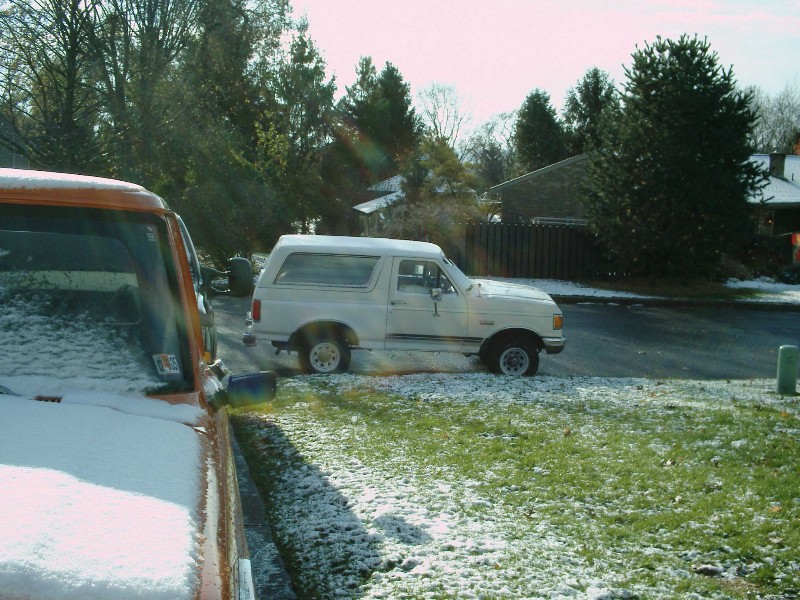 White Ford Bronco SUV parked on a snowy street, viewed from the side past an orange truck's snow-covered hood.