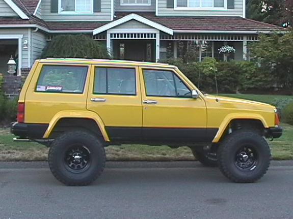 Side view of a lifted yellow Jeep Cherokee XJ with black steel wheels and big tires parked by a house