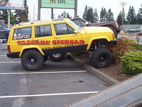Lifted yellow Jeep Cherokee with 'Cascade Offroad' lettering, front wheel on a curb in a parking lot