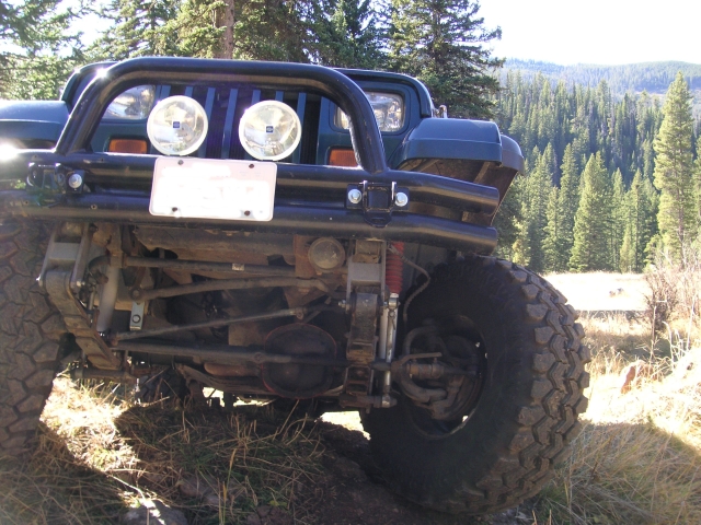 Dark green Jeep Wrangler front end with aftermarket bumper and off-road tires, parked on uneven terrain with a forest in the background