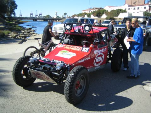 1990 Ford Bronco - red single-seat off-road race buggy numbered 92 with BFGoodrich and HDI sponsor decals sits on a waterfront lot as people inspect it.