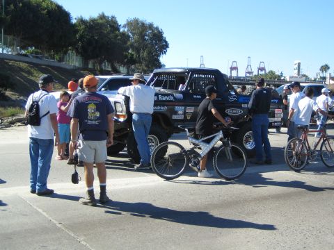 1990 Ford Bronco - Spectators and cyclists cluster around a dark caged off-road truck parked on a street, with shipping-port cranes visible in the distance.