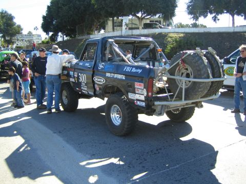 1990 Ford Bronco - black off-road race truck numbered 300 with a roll cage, knobby tires and rear-mounted spare sits on pavement as a crowd of spectators look…