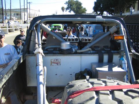 1990 Ford Bronco - View into the rear of an open off-road vehicle showing roll cage tubing, engine components and a turn-signal light, with spectators and oth…