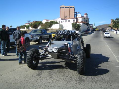 1990 Ford Bronco - silver off-road buggy with a roof light bar and long-travel suspension sits on a street, with pickups, spectators and a large hilltop build…
