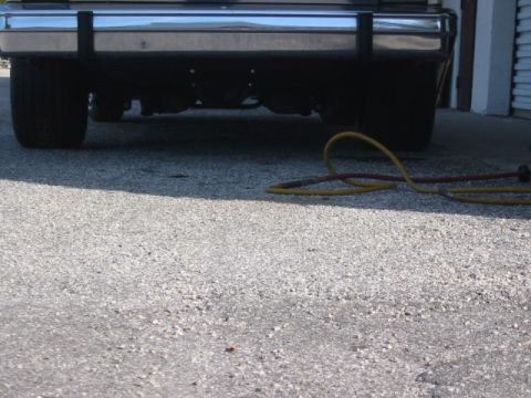 1985 Ford LTD Crown Victoria - A low-angle view of the rear of a vehicle, showing its bumper and tires, with a coiled hose on the asphalt.