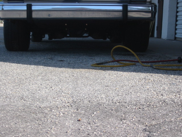 Car's chrome bumper with black protective strips, rear tires, and a yellow and red hose on the ground