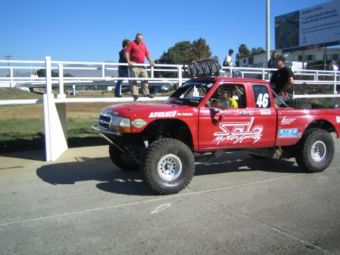 1990 Ford Bronco - red off-road race truck numbered 46 with roof light bar and 'Solo Motorsports' graphics sits on pavement near a white fence with spectators…