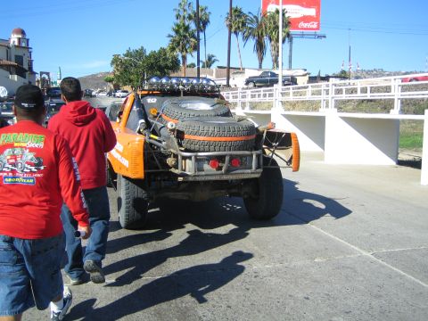 1990 Ford Bronco - Rear view of an orange off-road race truck carrying spare tires on a palm-lined street, with people in red shirts walking past and a Coca-C…