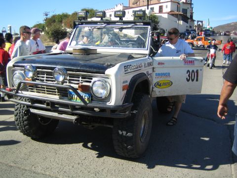 1990 Ford Bronco - early Ford Bronco off-road race vehicle numbered 301, white with a black hood and roof HID lights, surrounded by spectators on a street.