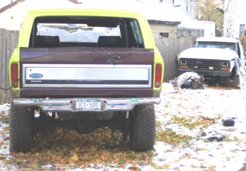 1978 Ford Bronco - A yellow and brown Ford Bronco is parked outside with a second white Ford truck in the background, both covered in a light dusting of snow.