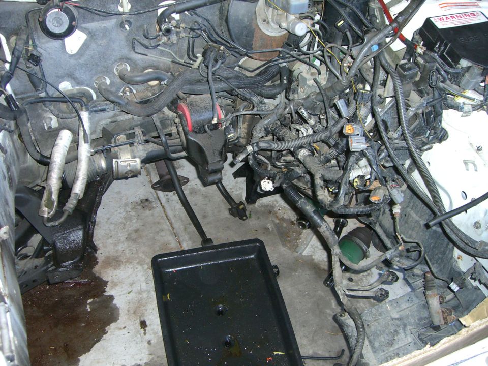 Engine bay with engine removed, showing wiring, hoses, and a drain pan with fluid