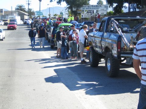 1990 Ford Bronco - Spectators stand along a sunlit street beside a row of staged off-road race vehicles, with a blue race pickup prominent in the foreground a…