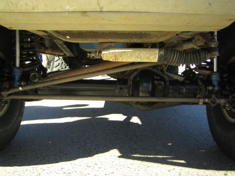 1990 Ford Bronco - Looking up under a vehicle at the front solid axle, coil springs, shock and steering tie-rod over a paved surface.