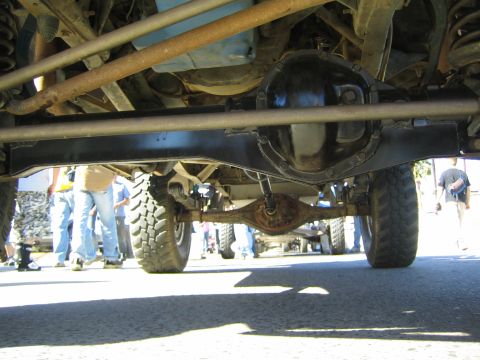 1990 Ford Bronco - Underbody view of an off-road truck showing the rear axle housing, driveshaft and knobby tires, with spectators' legs visible beyond.