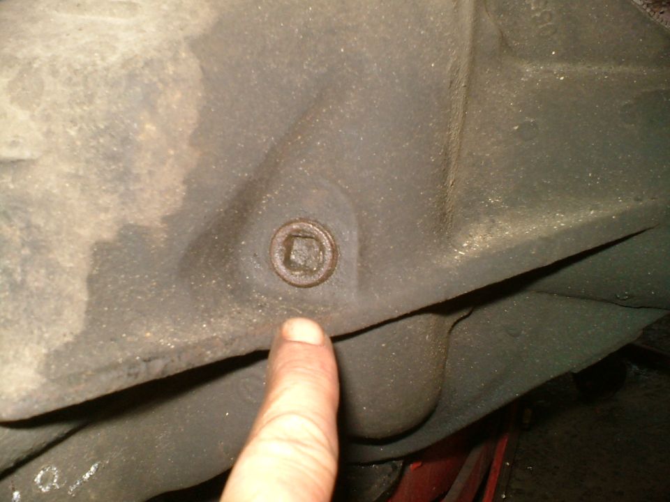 Close-up of a square-headed plug on a greasy metal housing, with a finger pointing to it