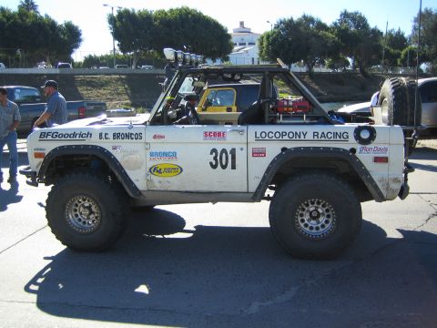 1990 Ford Bronco - Side profile of a white early Ford Bronco off-road racer numbered 301 with 'B.C. Broncos' and 'Locopony Racing' graphics and rear-mounted s…