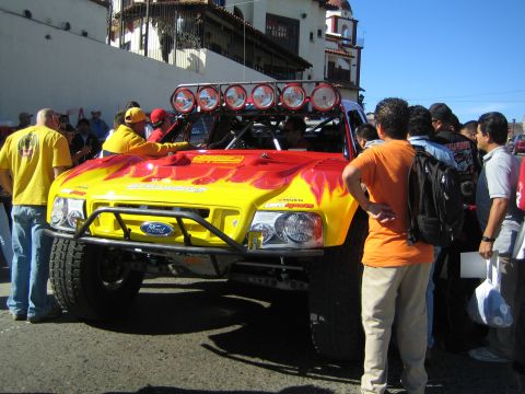 1990 Ford Bronco - yellow and red Ford off-road race truck with a row of roof lights and flame graphics is surrounded by a crowd in front of a building.