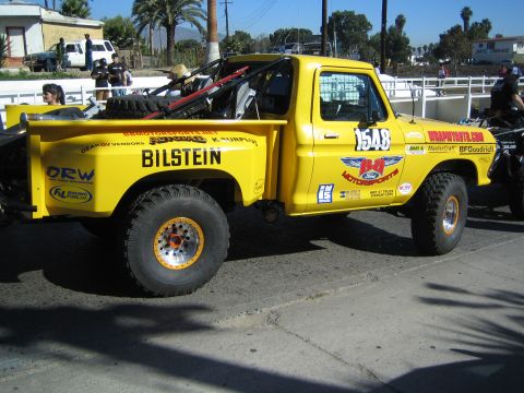 1990 Ford Bronco - yellow off-road race pickup numbered 1548 with 'Bilstein' and Ford Motorsports graphics sits on a street near a white fence and palm trees.