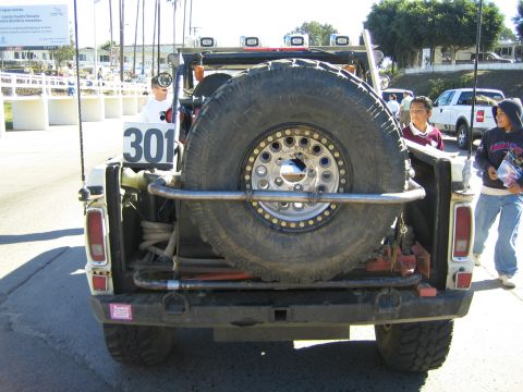 1990 Ford Bronco - Rear view of a white off-road race vehicle numbered 301 with a large beadlock spare tire mounted across the rear, two kids standing at righ…