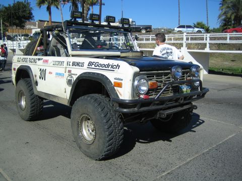 1990 Ford Bronco - white early Ford Bronco off-road racer numbered 301 with a black hood, roof HID lights and 'Old Horse Racing' graphics drives on pavement.