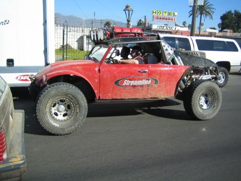 1990 Ford Bronco - red open-cockpit off-road Baja buggy with Streamline graphics is driven on a street past a box trailer and a white SUV.