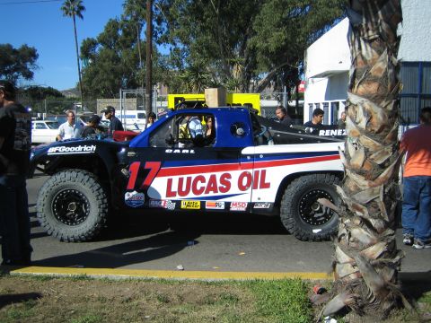 1990 Ford Bronco - blue-and-white off-road race truck numbered 17 with Lucas Oil and BFGoodrich branding sits among onlookers near a palm tree.