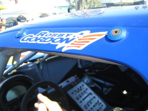 1990 Ford Bronco - blue race car body carries a Robby Gordon star-spangled decal above the windshield while a driver's hand rests on the wheel.