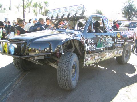 1990 Ford Bronco - black off-road race truck with a front roof light bar and Cabo Wabo and Yokohama sponsor decals sits surrounded by spectators.
