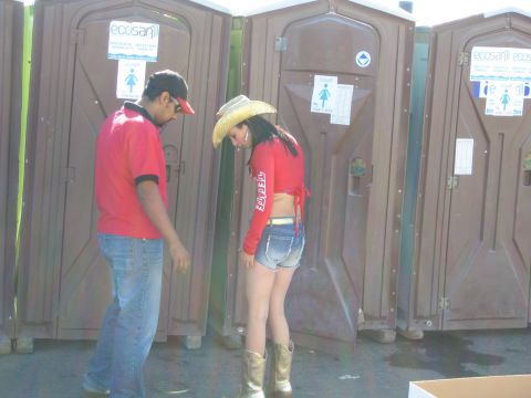 1990 Ford Bronco - man and a woman in red shirts stand looking down in front of a row of brown portable toilets at an outdoor event.