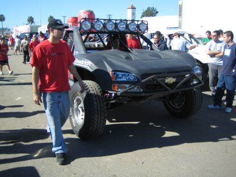 1990 Ford Bronco - silver-and-black Chevrolet off-road race truck with a roof light bar is pushed through a crowd as a man in red walks alongside.