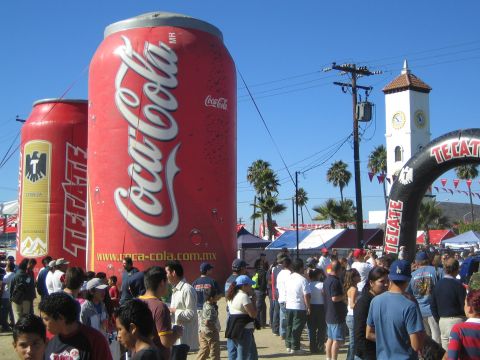 1990 Ford Bronco - Crowds gather beneath giant inflatable Coca-Cola and Tecate beverage cans at a daytime outdoor event with a clock tower behind.