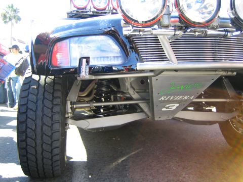 1990 Ford Bronco - Detailed view of a blue off-road racer's front suspension showing coilover shock, control arms and chrome lighting, marked Riviera 3.