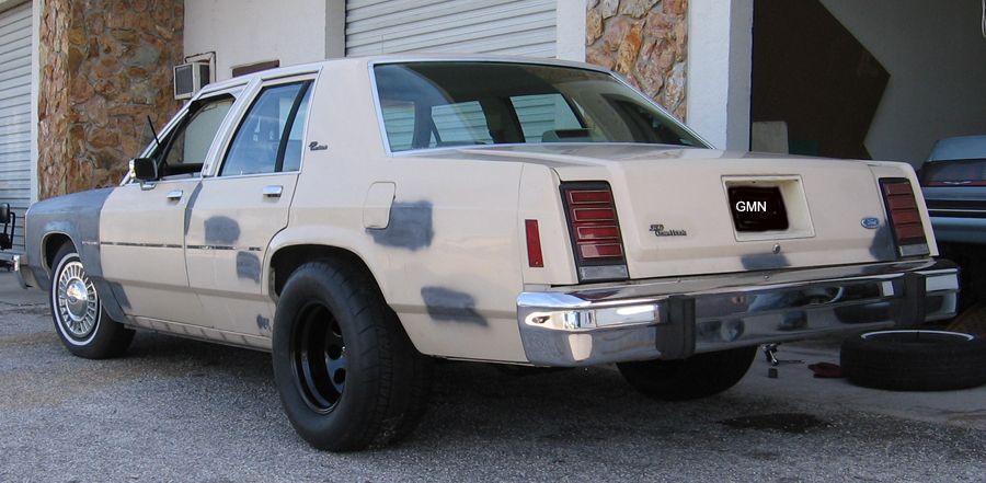 Rear view of a beige Ford sedan with primer patches on the body, black wheels, and a license plate that reads GMN