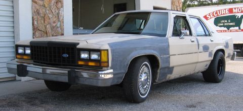 1985 Ford LTD Crown Victoria - A beige sedan with primer-gray body panels is parked in front of a building and a moving truck.
