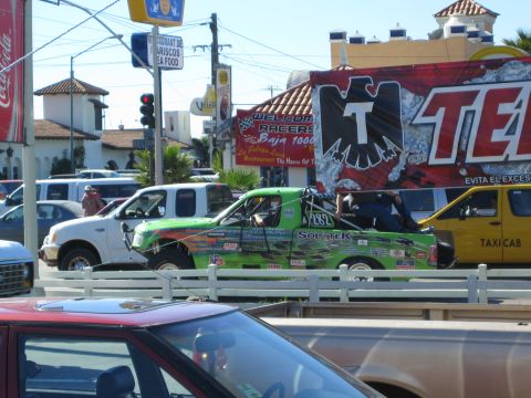 1990 Ford Bronco - green tube-frame off-road race truck rides on a trailer through a street lined with vehicles and large Tecate and Coca-Cola banners.