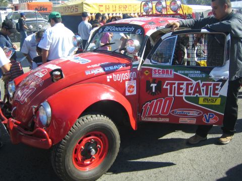 1990 Ford Bronco - red Baja-prepped VW Beetle race car numbered 1100 with Tecate and BFGoodrich decals sits with its door held open at an event.