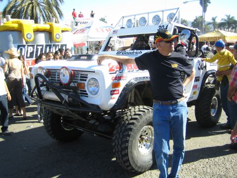 1990 Ford Bronco - man in a black shirt and cap leans against a white number 304 off-road Bronco-style race truck amid a festival crowd.