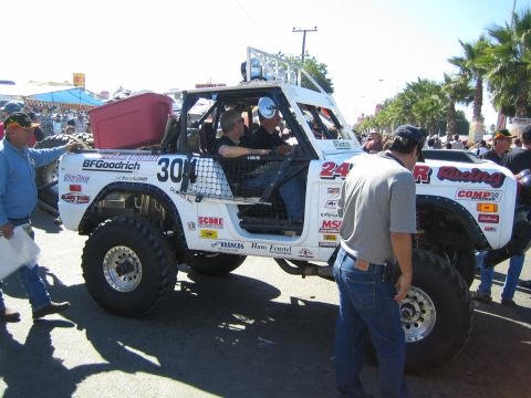 1990 Ford Bronco - Two occupants sit in a white number 304 tube-frame off-road race truck as bystanders gather around it on a sunny street.