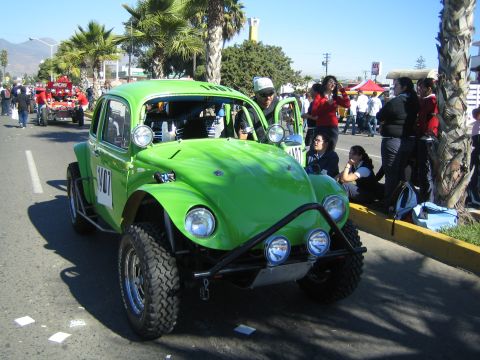 1990 Ford Bronco - bright green Baja-prepped VW Beetle off-road car with extra lights and big tires sits on a palm-lined street as people gather.