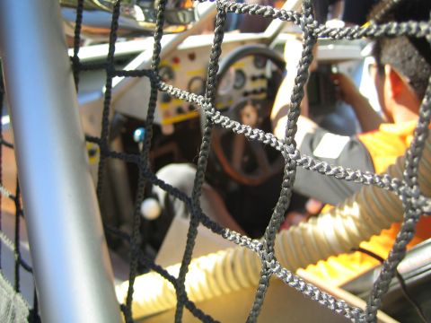 1990 Ford Bronco - Looking through a black safety window net into a race car cockpit showing the steering wheel, dash gauges and a person beside it.