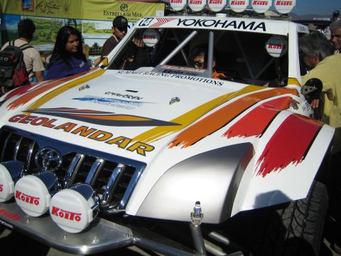 1990 Ford Bronco - white off-road race car with red and orange Geolandar and Yokohama livery and capped Koito lights is inspected by spectators.