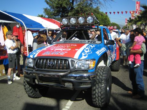 1990 Ford Bronco - red, white and blue Toyota Tacoma off-road race truck numbered 720 with roof lights drives down a crowded festival street.