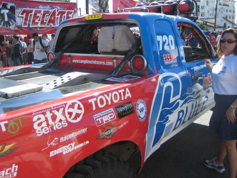 1990 Ford Bronco - red and blue Toyota off-road race truck numbered 720 covered in sponsor decals, with a woman standing beside it at an outdoor event.