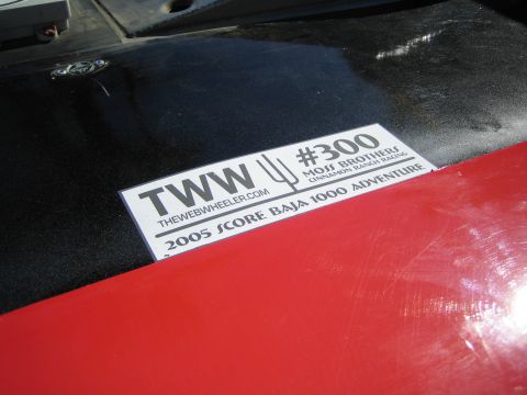 1990 Ford Bronco - Close-up of a 'TWW #300 Moss Brothers 2005 Score Baja 1000 Adventure' sponsor decal on a glossy black and red vehicle panel.