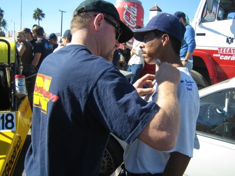1990 Ford Bronco - Two men in caps and sunglasses converse closely amid a crowd at an outdoor event, with a yellow race vehicle and Coca-Cola sign behind them.